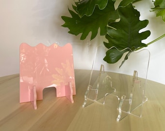 May include: Two phone stands, one pink and one clear, on a wooden surface. The pink stand has a wavy design and the clear stand has a simple design.
