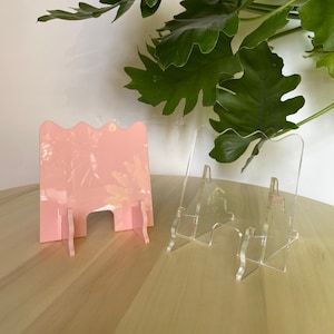 May include: Two phone stands, one pink and one clear, on a wooden surface. The pink stand has a wavy design and the clear stand has a simple design.