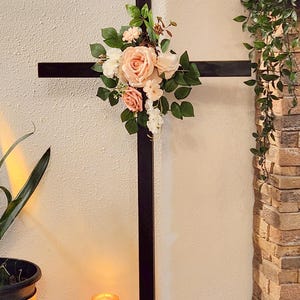 May include: A tall, dark brown wooden cross standing on a square base. The center of the cross is adorned with a cluster of artificial flowers in shades of peach, cream, and white, accented with green leaves and small sprigs of greenery.
