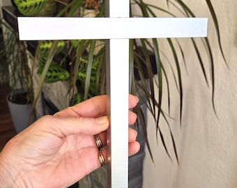 May include: A silver-colored cross with a smooth, matte finish and a simple, clean-lined design. The object features a vertical bar intersected by a shorter horizontal bar, forming a standard cross shape with flat, uniform edges.