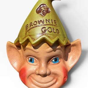May include: A vintage ceramic wall hanging featuring a smiling elf face with blue eyes and rosy cheeks. The elf wears a green hat with the words "Brownie Gold" printed on it. The design is whimsical and decorative.
