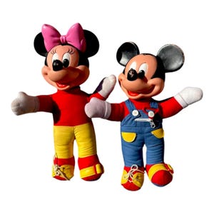 May include: Two vintage plush Disney characters, Mickey and Minnie Mouse, stand side-by-side. Minnie wears a pink bow, red top, yellow pants, and red shoes. Mickey wears blue overalls, a red top, and red shoes.