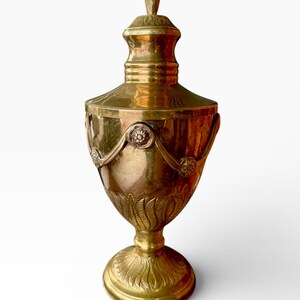 May include: A gold-colored urn with a lid, featuring ornate detailing. The urn has a bulbous body, a pedestal base, and a decorative finial on top. The design includes floral and ribbon motifs.