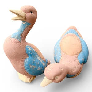 May include: Two ceramic duck figurines. The ducks are painted in a distressed style with a pinkish-brown body, light blue accents, and off-white beaks. One duck is upright, and the other is lying down.