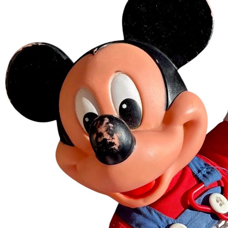 May include: Close-up of a vintage Mickey Mouse figurine. The figurine features a peach-colored face, black ears, and a black nose with some wear. Mickey is wearing a red top and blue overalls. The image is set against a white background.