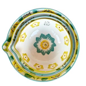 May include: A set of four ceramic measuring bowls. The bowls are white with yellow and teal floral designs. The rims are teal and yellow. The largest bowl has the word "ONE" written on it. The smallest bowl has "1/4 CUP" written on it.