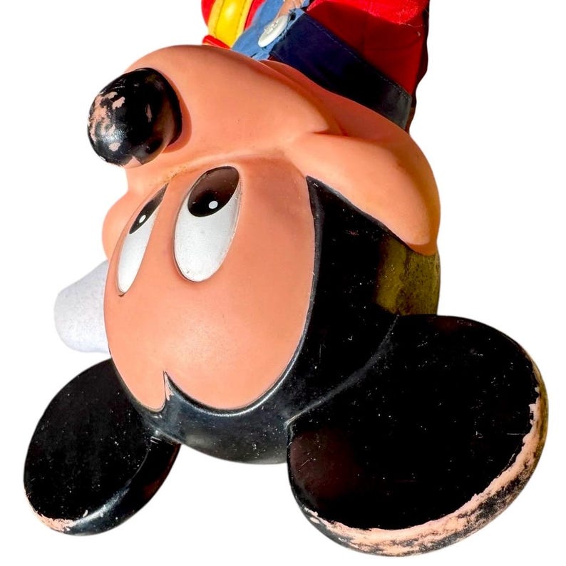 May include: A vintage Mickey Mouse figurine, featuring a smiling face with large, oval eyes and black ears. The figure has a red, blue, and yellow hat. The figurine is made of a glossy material with some wear and tear.