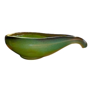 May include: A green ceramic serving dish with a unique, elongated shape and a curved handle. The dish has a speckled glaze and a glossy finish.