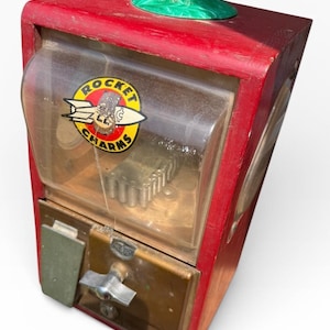May include: Vintage red wooden gumball machine with a clear plastic front. The machine features a "Rocket Charms" sticker and a green plastic top. The coin slot and dispensing lever are visible on the front.