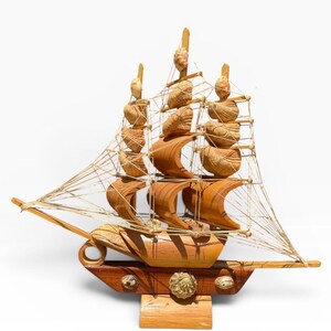 May include: A handcrafted wooden model ship with three masts and detailed sails. The ship is made of light and dark brown wood, with intricate carvings and rope detailing. The model sits on a wooden base with decorative accents.