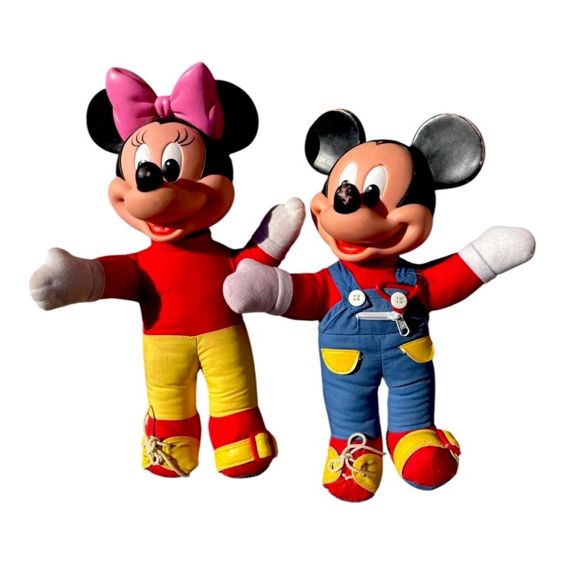 May include: Two plush Disney character dolls, Mickey and Minnie Mouse, stand side-by-side. Minnie wears a pink bow, red top, and yellow pants. Mickey wears blue overalls and a red top. Both have white gloves and red shoes.