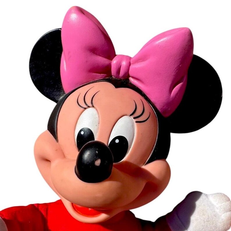 May include: A close-up of a Minnie Mouse figurine. The figurine has a pink bow, black ears, and a red top. The face is peach-colored with black eyes and a black nose. The background is white.