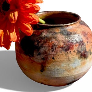 May include: A handcrafted, round ceramic vase with a textured, mottled surface in shades of brown, orange, and black. A vibrant orange flower with green stem is placed inside the vase, creating a striking contrast.