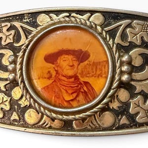May include: A rectangular metal belt buckle featuring a central circular portrait of a person wearing a wide-brimmed hat and a neckerchief. The surrounding frame is decorated with embossed oak leaf and acorn motifs in a dark, aged gold finish.
