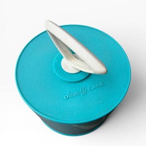 May include: A teal-colored Disk-Go Case with a white handle. The case is round with a dark gray band around the side. The text "Disk-Go Case" is printed on the front of the case.