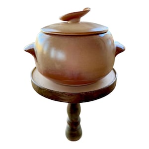 May include: A brown ceramic soup tureen with a lid and two handles, sitting on a wooden pedestal.