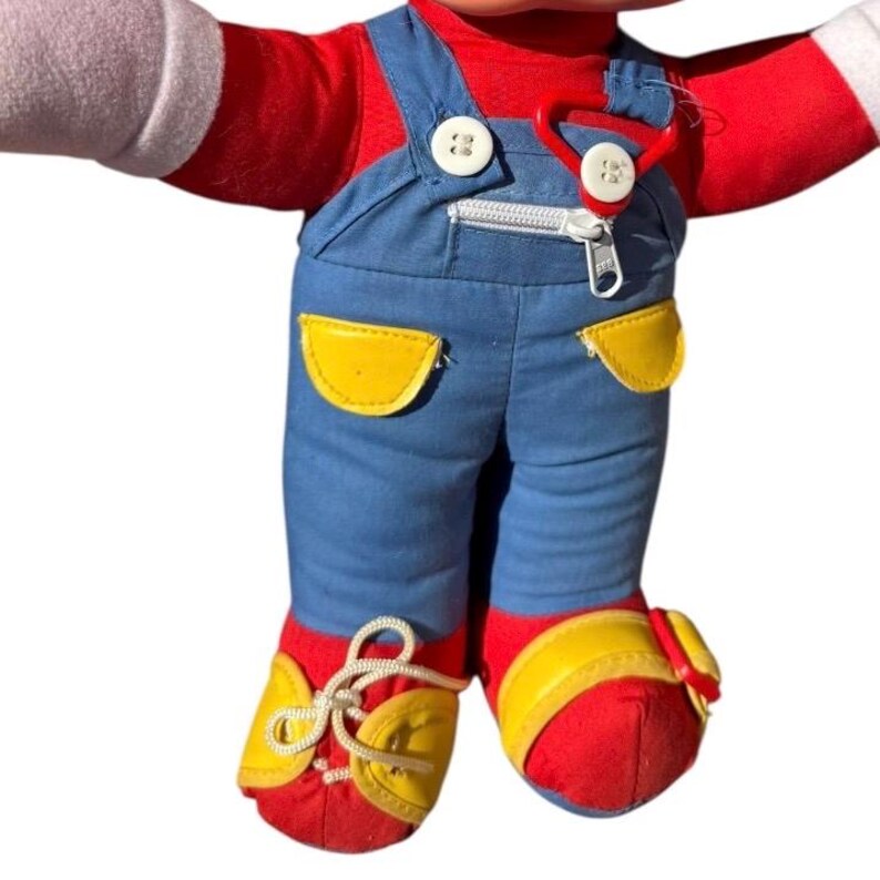 May include: A plush doll wearing a red shirt, blue overalls with yellow pockets, and red shoes with yellow accents. The doll has white gloves and a red plastic clasp attached to the overalls.