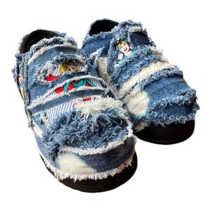 May include: A pair of denim slip-on shoes with frayed edges. The shoes feature a comic book style print of female superheroes.