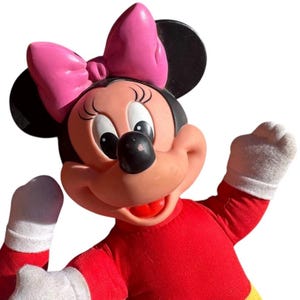 May include: A Minnie Mouse plush doll with a pink bow, black ears, and a red dress. The doll has white gloves and a smiling expression. The background is white.