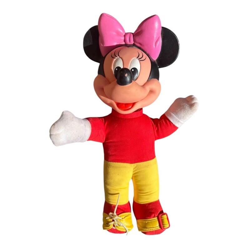 May include: A vintage Minnie Mouse plush doll. The doll has a red top, yellow pants, and red shoes. Minnie's signature pink bow sits atop her black ears. Her white gloves are outstretched in a welcoming gesture. The doll is a classic Disney collectible.