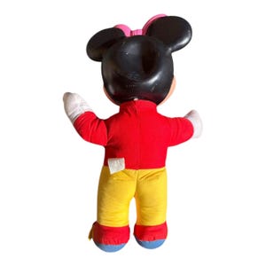 May include: A vintage Minnie Mouse plush doll, viewed from the back. The doll features a black head with large ears and a pink bow. It wears a red top, yellow pants, white gloves, and blue shoes. The doll is a classic Disney character.