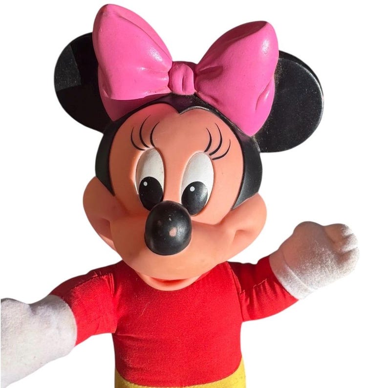May include: A Minnie Mouse plush toy with a pink bow, black ears, and a red shirt. The toy has white gloves and a yellow skirt. The character's face is peach-colored with black eyes and a black nose.