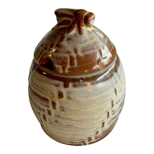 May include: A ceramic honey pot shaped like a beehive with a bee on top. The pot is glazed in brown and yellow with a textured surface.