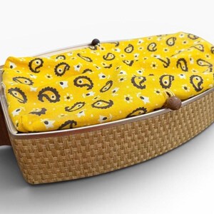 May include: A vintage, boat-shaped basket with a woven, light brown exterior. The interior features a bright yellow fabric with a paisley pattern in black and white. The basket has a metal frame and wooden accents.