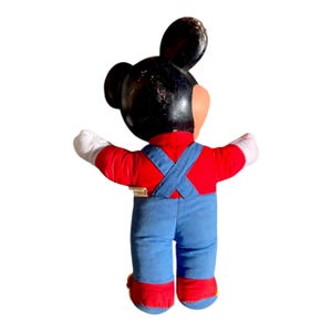 May include: Vintage Mickey Mouse plush toy, viewed from the back. Mickey is wearing a red shirt, blue overalls, and red and yellow shoes. His black head has visible wear, and his white gloved hands are outstretched.
