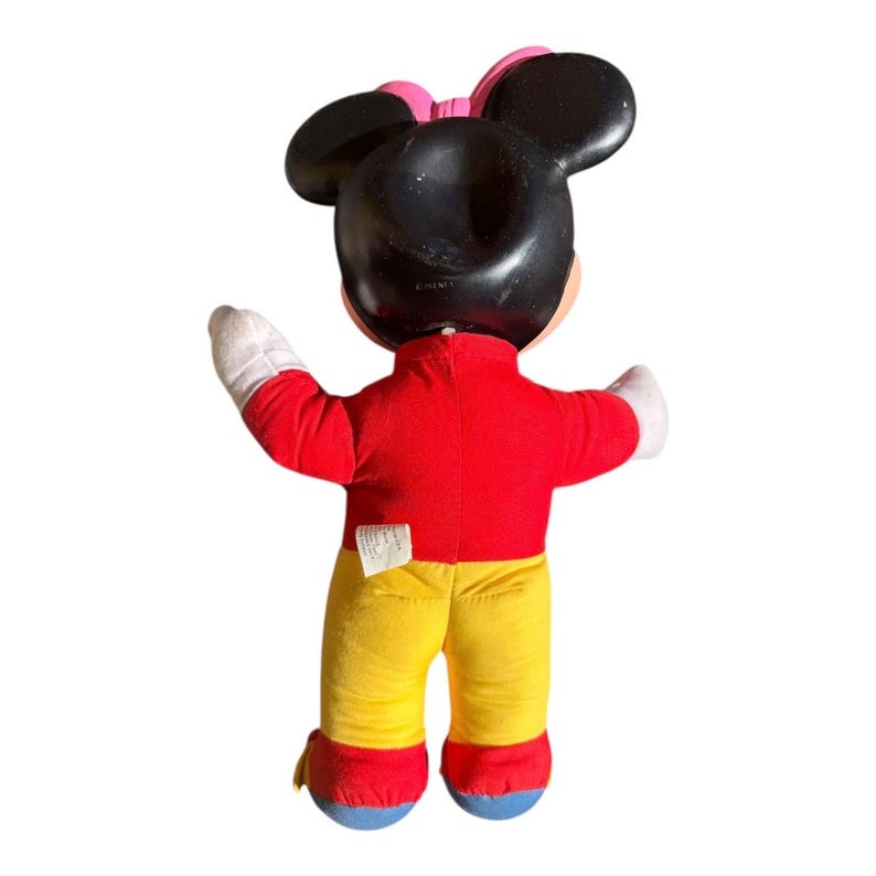 May include: A plush Minnie Mouse doll, viewed from the back, with a black head and ears, a pink bow, and a red top. The doll has yellow pants, white gloves, and red and blue shoes. The doll's arms are outstretched.