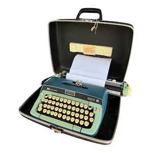 May include: A vintage blue and green typewriter with cream-colored keys sits inside a black carrying case. The typewriter has a paper inserted and the words "Galaxie Twelve" are visible on the front.