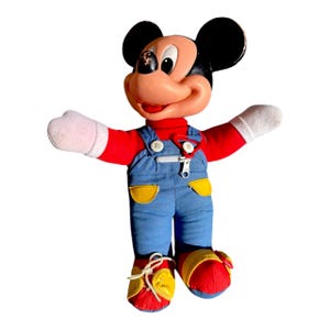 May include: Vintage Mickey Mouse plush doll. The doll has a red shirt, blue overalls with yellow pockets, and red and yellow shoes. Mickey's face is tan with black ears and a red mouth. The doll's arms are outstretched.