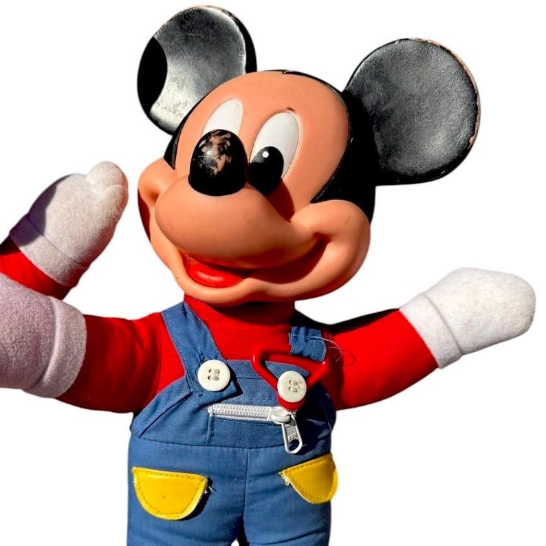 May include: A vintage Mickey Mouse plush doll. The doll has a red shirt, blue overalls with yellow pockets, and white gloves. Mickey's face is peach-colored with black eyes and a black nose. The ears are black.