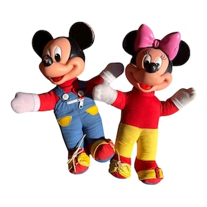 May include: Two vintage Mickey and Minnie Mouse plush dolls. Mickey wears blue overalls, a red shirt, and yellow shoes. Minnie wears a red shirt, yellow pants, a pink bow, and yellow shoes. Both dolls have black ears and white gloves.