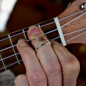 May include: A close-up of a hand wearing a silver finger pick with a red gemstone on a ukulele. The ukulele has a brown wooden body and white nylon strings.