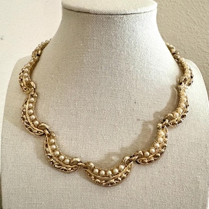 May include: A gold-toned necklace with a scalloped design, adorned with small, round, white faux pearls. The necklace is displayed on a white mannequin bust, showcasing its intricate details and vintage style.
