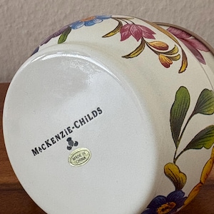 May include: A ceramic vase with a cream-colored base and floral designs in shades of purple, blue, yellow, and green. The bottom of the vase features the text "MACKENZIE-CHILDS" and a "Made in China" sticker.