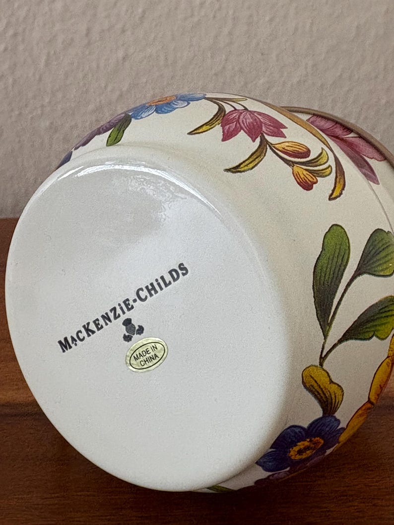 May include: A ceramic vase with a cream-colored base and floral designs in shades of purple, blue, yellow, and green. The bottom of the vase features the text "MACKENZIE-CHILDS" and a "Made in China" sticker.