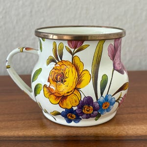 May include: A white enamel mug with a floral design featuring yellow, purple, and blue flowers. The mug has a bronze rim and handle. The floral pattern is set against a white background.