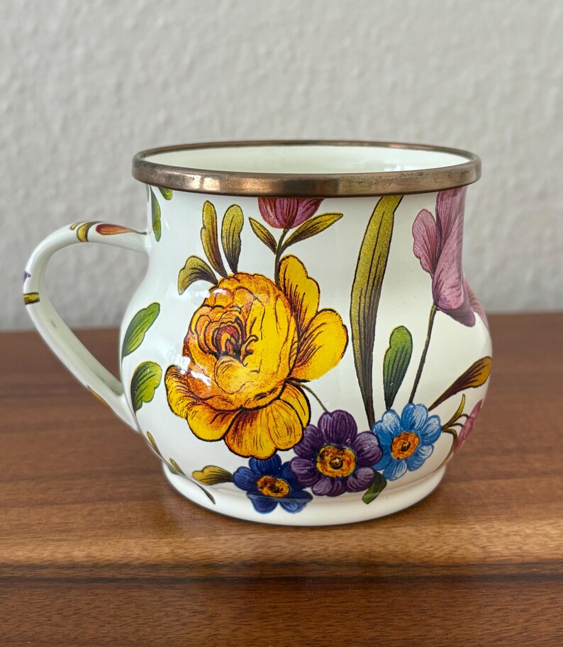 May include: A white enamel mug with a floral design featuring yellow, purple, and blue flowers. The mug has a bronze rim and handle. The floral pattern is set against a white background.