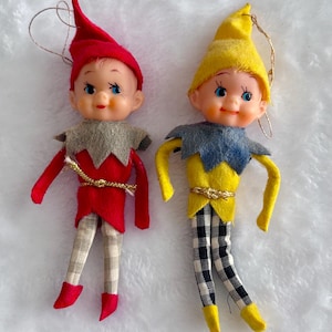 May include: Two vintage elf ornaments. One elf wears a red outfit with a matching hat, gray collar, and plaid pants. The other elf has a yellow outfit, a blue collar, and black and white checkered pants. Both have blue eyes and are smiling.