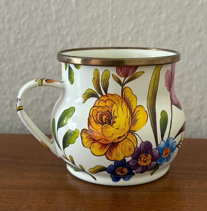 May include: White enamel mug with a floral design featuring a large yellow rose, purple, and blue flowers. The mug has a gold rim and handle, and the floral pattern extends around the body of the mug.