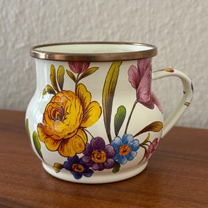 May include: A white enamel mug with a floral design featuring yellow, purple, and blue flowers. The mug has a brass rim and handle, and the flowers are painted on the exterior. The mug is sitting on a wooden surface.