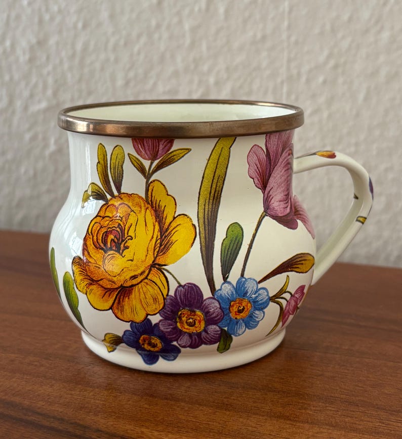 May include: A white enamel mug with a floral design featuring yellow, purple, and blue flowers. The mug has a brass rim and handle, and the flowers are painted on the exterior. The mug is sitting on a wooden surface.