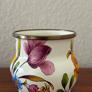 May include: A white ceramic pot with a bronze rim and floral designs. The pot features purple, blue, and yellow flowers with green leaves. The pot is sitting on a wooden surface.