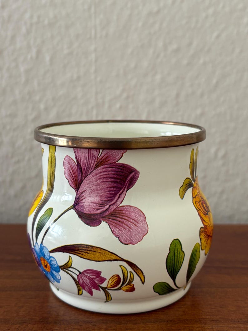 May include: A white ceramic pot with a bronze rim and floral designs. The pot features purple, blue, and yellow flowers with green leaves. The pot is sitting on a wooden surface.