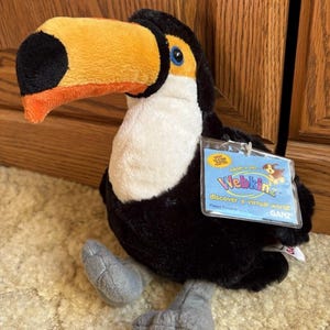 May include: A plush toucan toy with a large orange and yellow beak, black body, white chest, and gray feet. The toy has a Webkinz tag attached. The toucan is sitting on a beige carpet.