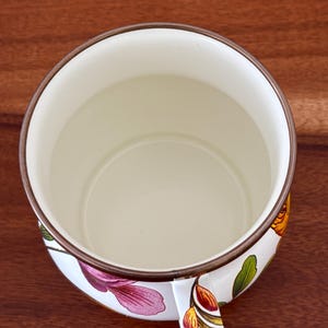 May include: A white enamel mug with a floral design featuring pink, yellow, and green flowers. The mug has a brown rim and handle, and sits on a dark wood surface. The interior of the mug is off-white.