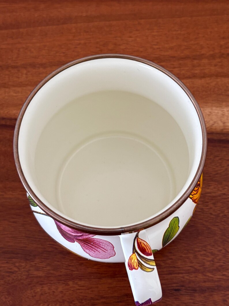 May include: A white enamel mug with a floral design featuring pink, yellow, and green flowers. The mug has a brown rim and handle, and sits on a dark wood surface. The interior of the mug is off-white.