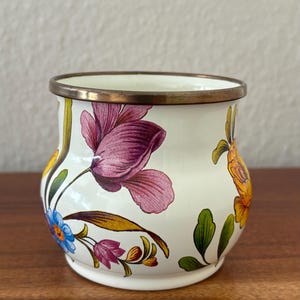 May include: A small, white ceramic vase with a floral design. The vase features a brass rim and is adorned with purple, blue, and yellow flowers with green leaves. The flowers are painted on the exterior of the vase.